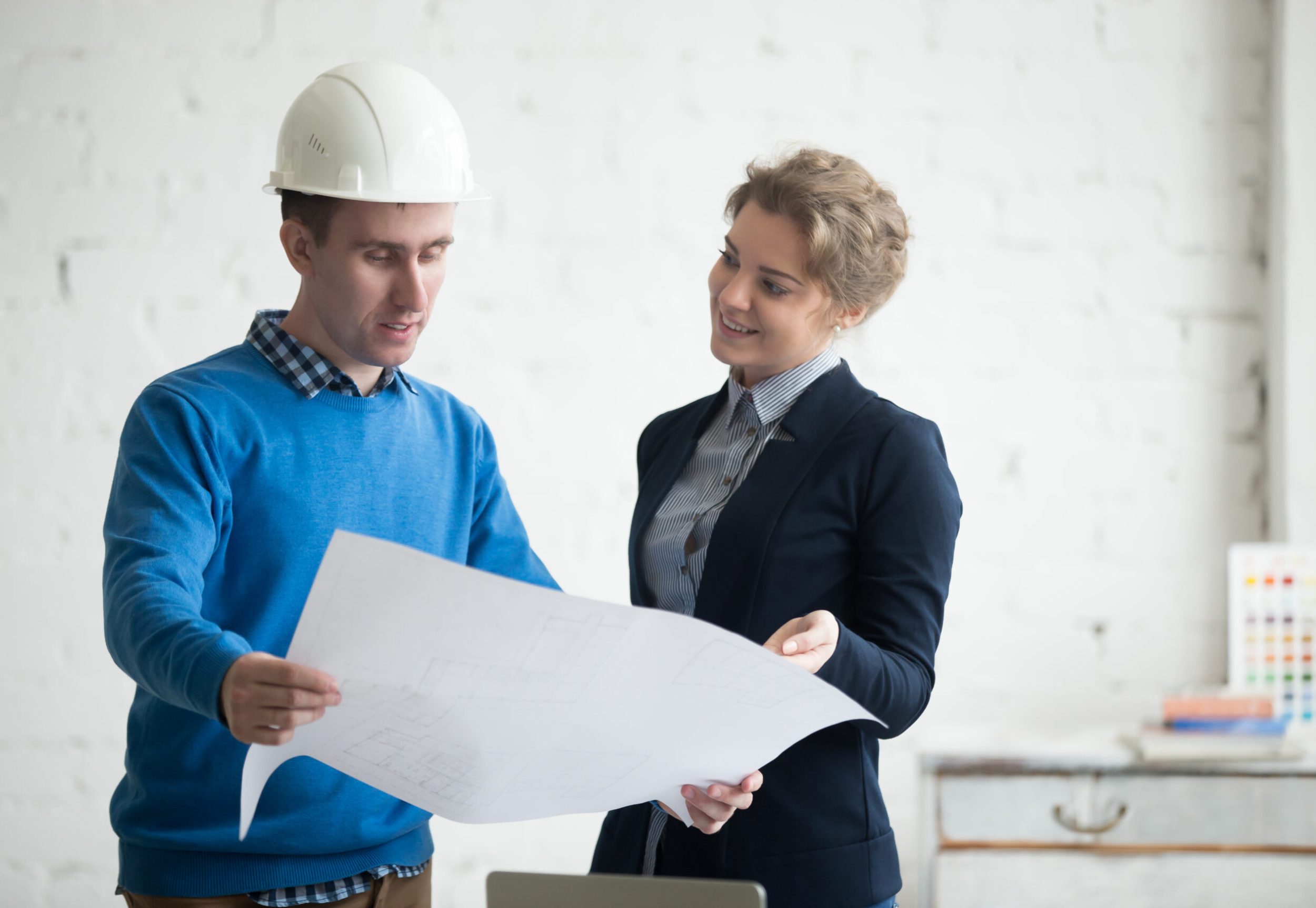 Two young colleagues at meeting looking at project concept, holding blueprints, planning work. Business discussion with construction engineer in hardhat in architectural agency office. Interior shot